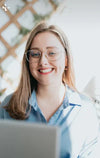 Smiling woman with glasses in blue shirt working on laptop in bright indoor setting